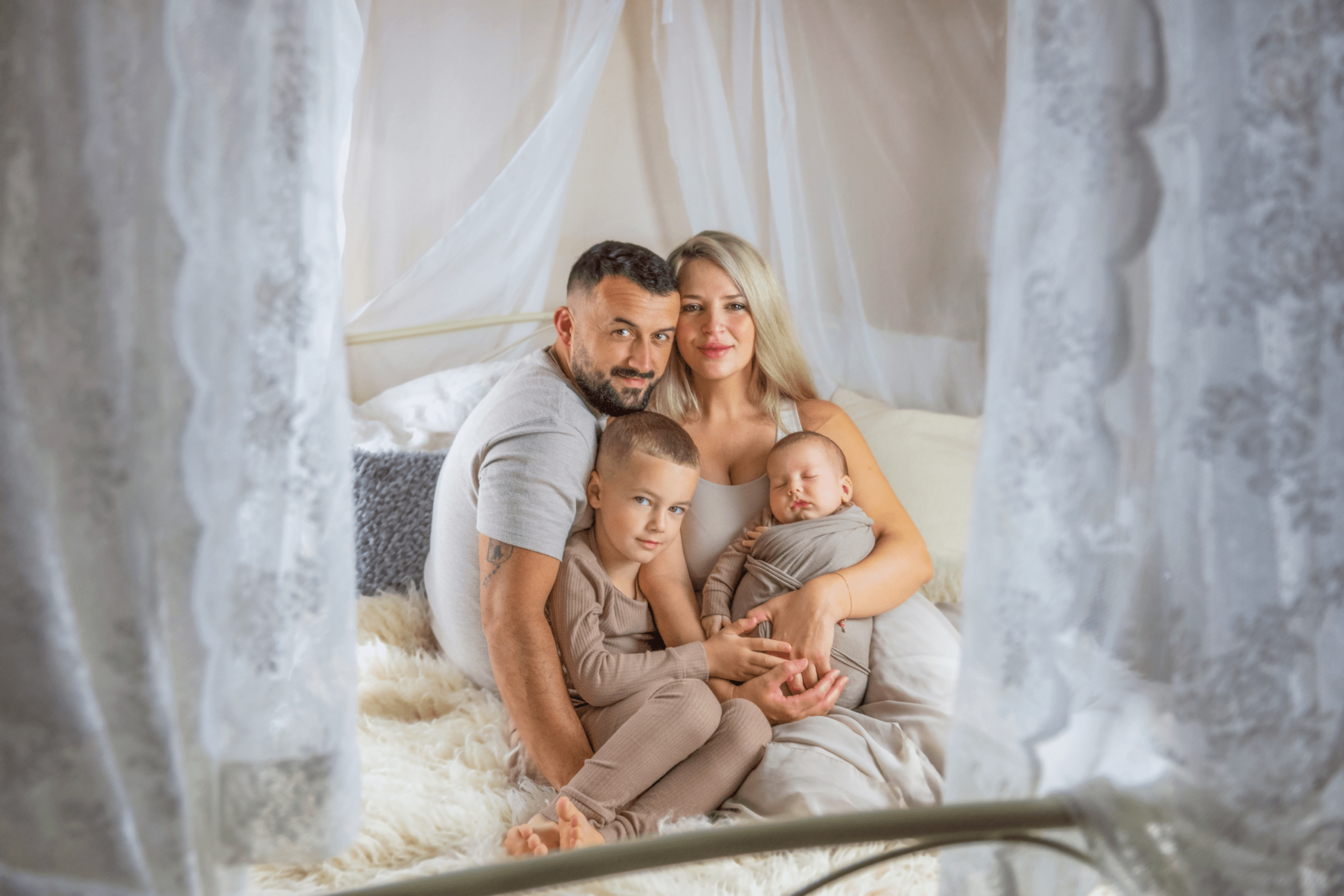 Eine Familie mit zwei Kindern sitz auf einem Himmelbett und schaut zur Fotografin. Im Vordergrund sind sind rechts und links helle Vorhänge zu erkennen.