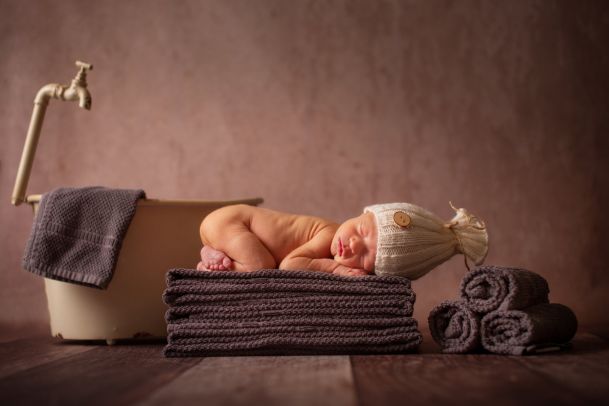 Ein kleines Baby mit weißer Mütze liegt  auf einem Stapel brauner Handtücher und schläft. Im Hintergrund eine kleine Badewanne.