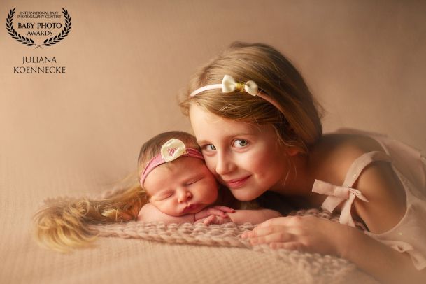 Ein kleine Baby lieft schlafend auf einer cremefarbenen Decke mit Blick zum Betrachter. Daneben schmiegt sich von rechts kommend, die größere Schwester an das Baby. Sie trägt einen kleinen Haarreifen.