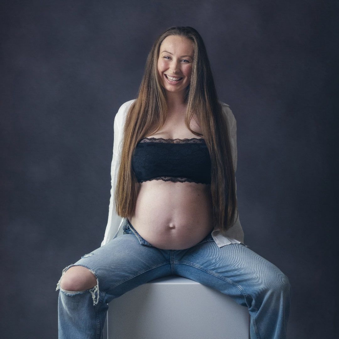 Das Bild zeigt eine schwangere Frau im Rahmen des Babybauch Shootings. Sie sitzt, lacht, hat den Bauch frei und trägt eine Jeanshose.