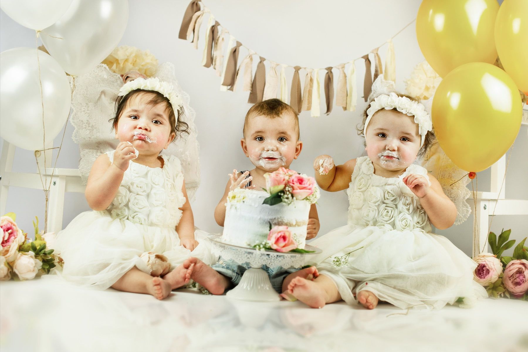 Three babies are sitting next to each other, a cake in the foreground, everything is decorated in white and pastel yellow for the birthday. The mouths are already smeared with cake.