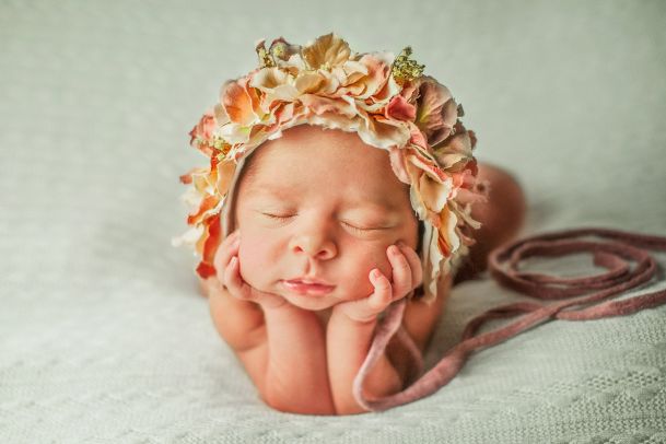 A sleeping baby, with a wreath of flowers on her head, lying in prone position resting her head on her hands and forearms.