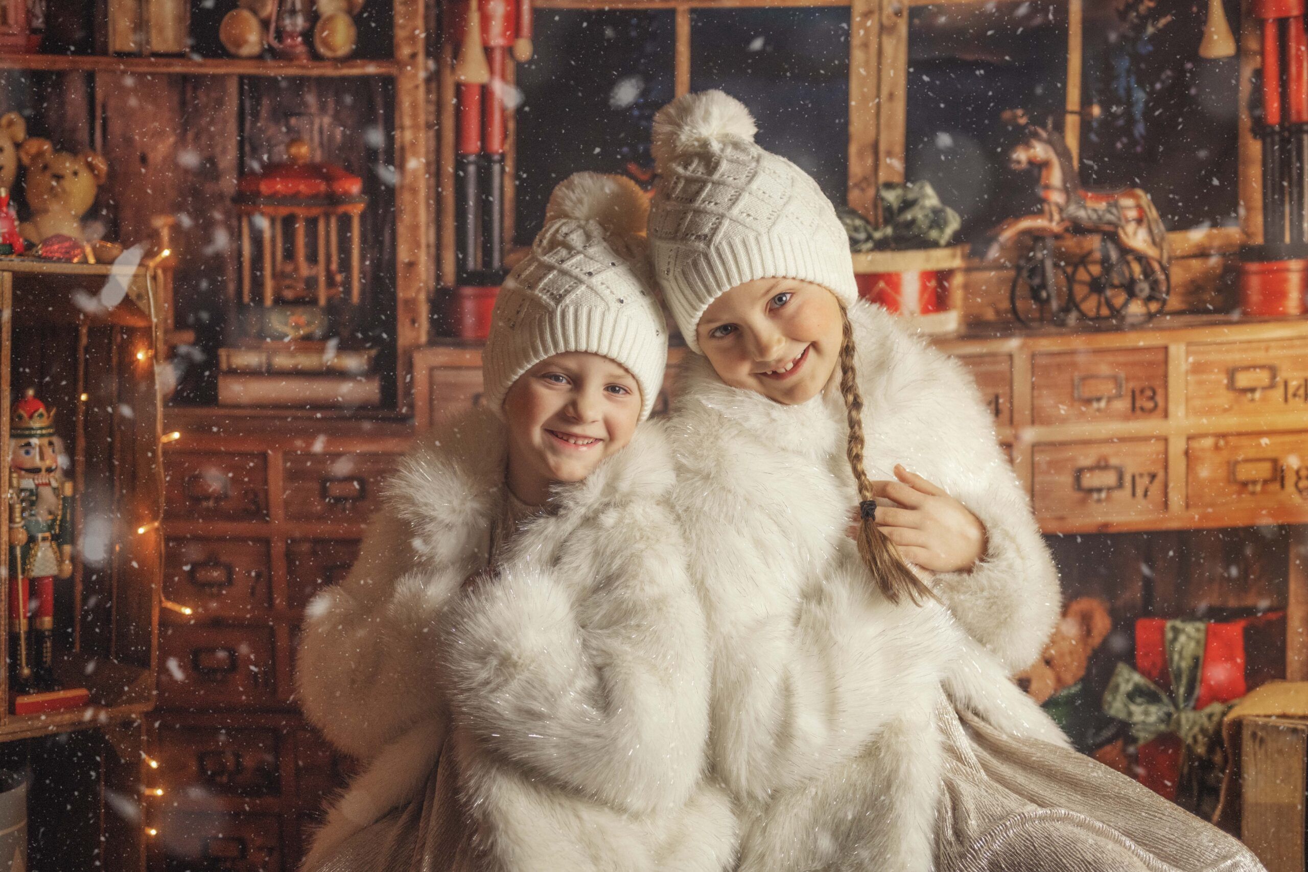 Zwei fröhliche Kinder in weißen Wintermänteln und Mützen posieren lachend in einer weihnachtlich dekorierten Stube.