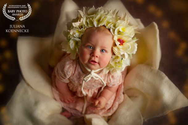 Ein kleines Baby sitzt in einer dekorativen Blume, hat einen Blumenkranz auf dem Kopf und schaut lächelnd nach oben, direkt zum Betrachter.