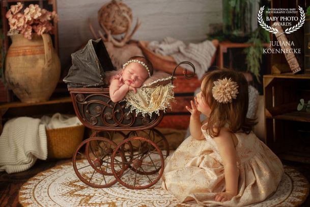 Ein Baby liegt schlafend in einem alten Kinderwagen aus dem 18. Jahrhunder. Dabei lehnt es sich etwas aus dem Kinderwagen. Auf dem Boden sitzt die ältere Schwester und schaut zu dem Baby.