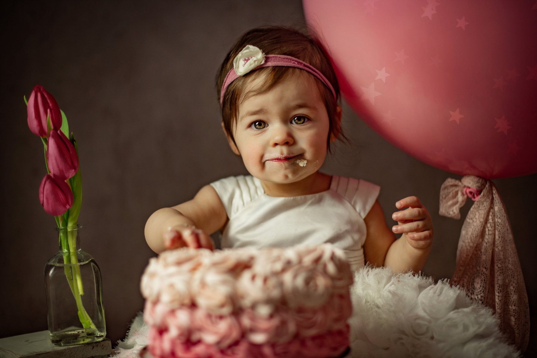 Baby with a pink headband are in front of a pink cake with a large pink balloon. The mouth is already a bit smeared with cake and cream.