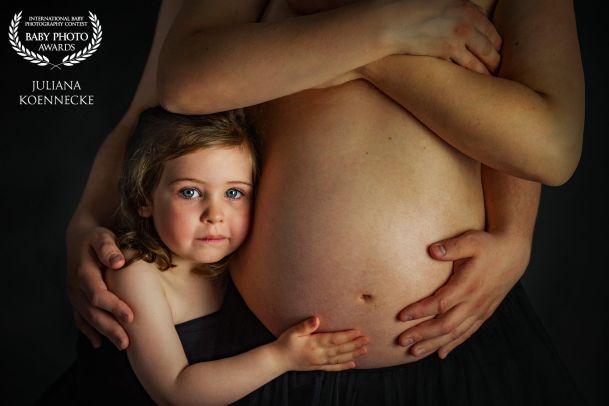 Man sieht den nackten Bauch einer schwangeren Frau. Daneben steht dicht, angeschmiegt ein kleines Mädchen und hält den Bauch. Der Mann umart die schwangere Frau von hinten. Zu sehen ist fast nur nackte Haut und ein schwarzer Hintergrund.