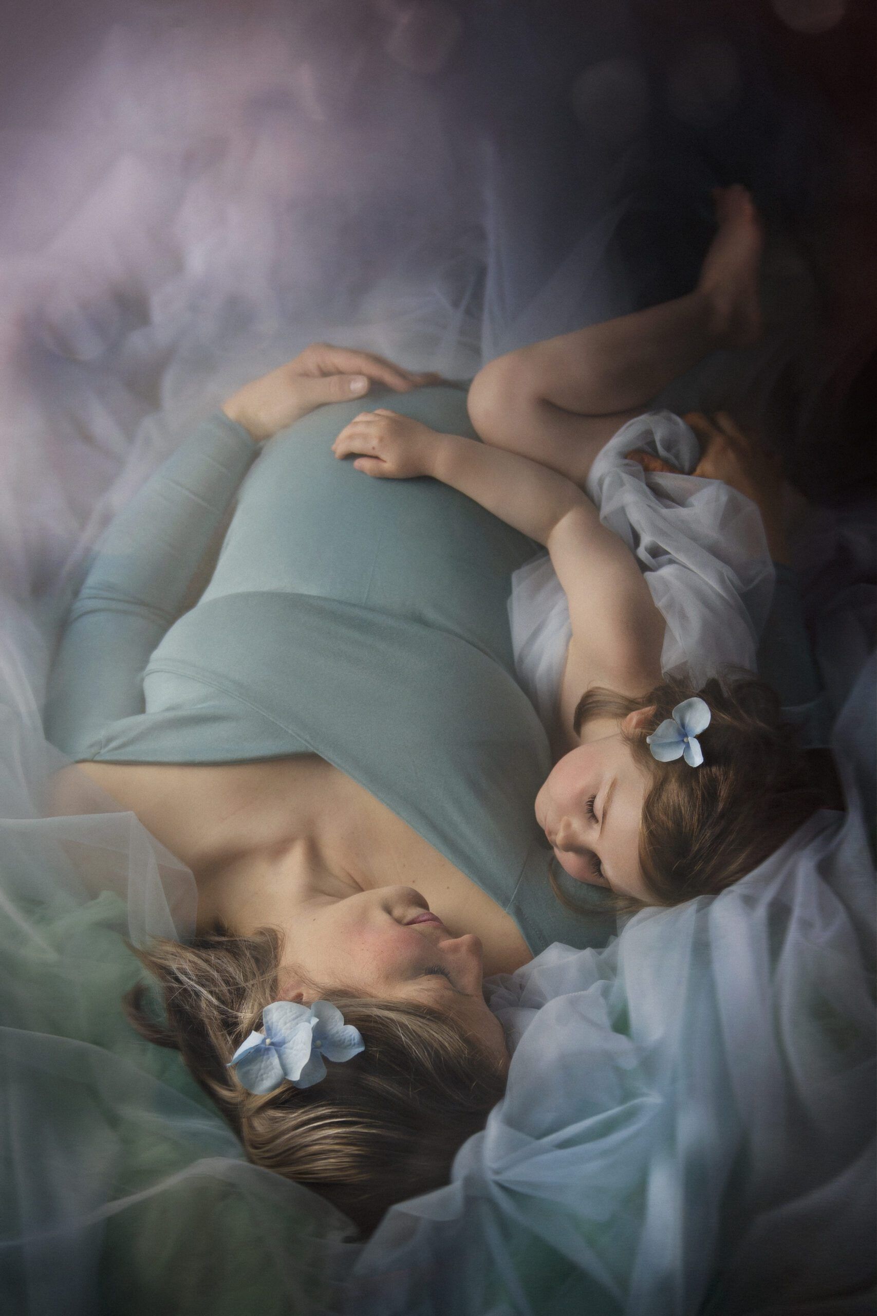 A pregnant woman in a pastel blue dress lies on her back and looks at her daughter, who is lying on her side to her right. Both hold the woman's belly.