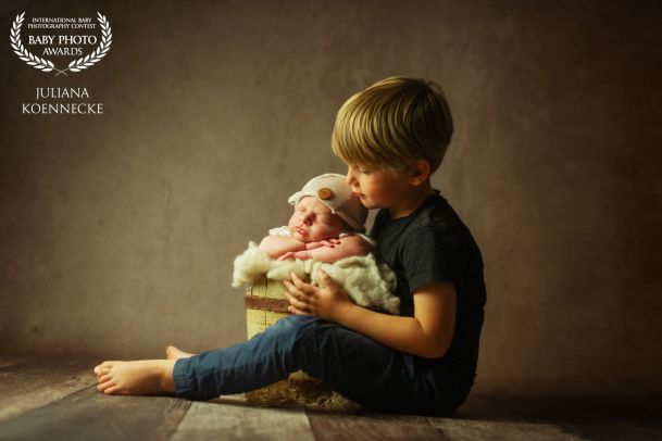 Ein kleiner Junge sitzt auf dem Fussboden und hat zwischen den Beinen einen Eimer stehen. In diesem Eimer sitzt ein Neugeborenes Baby, eingekuschelt in eine Decke, und schläft.