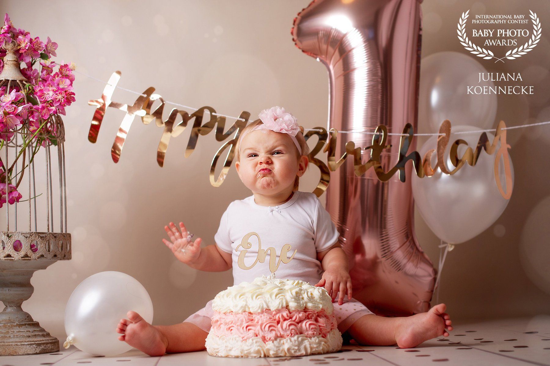 A baby sits on the floor with his legs apart. Right in front of the baby is a white and pink cream cake. Behind it a big 1 as a balloon and flower decoration.