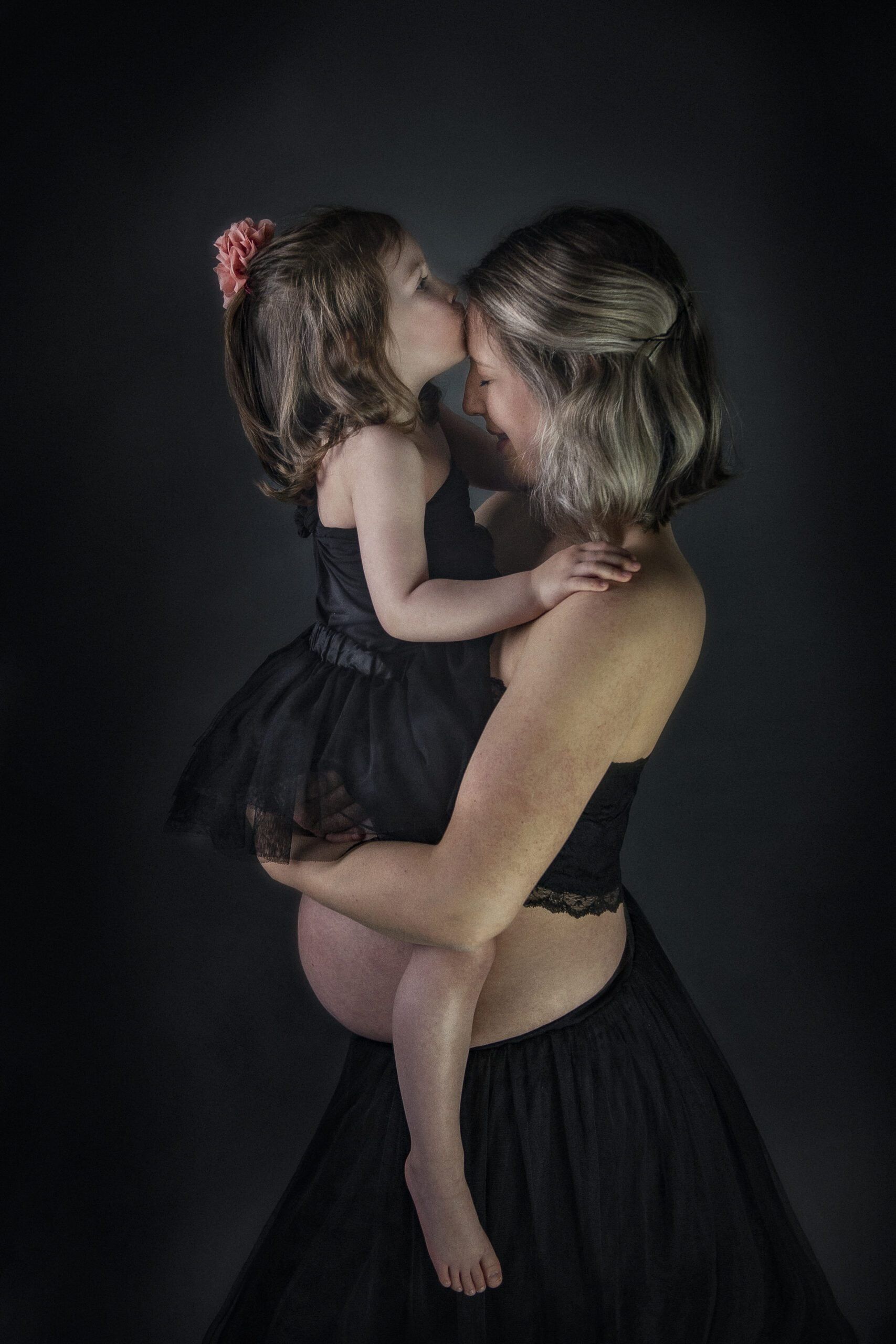 A pregnant woman in front of a dark background, photographed from the side. She wears a black skirt and a black top, arms and stomach are free. In her arms she is holding a young girl who is also wearing a black dress.