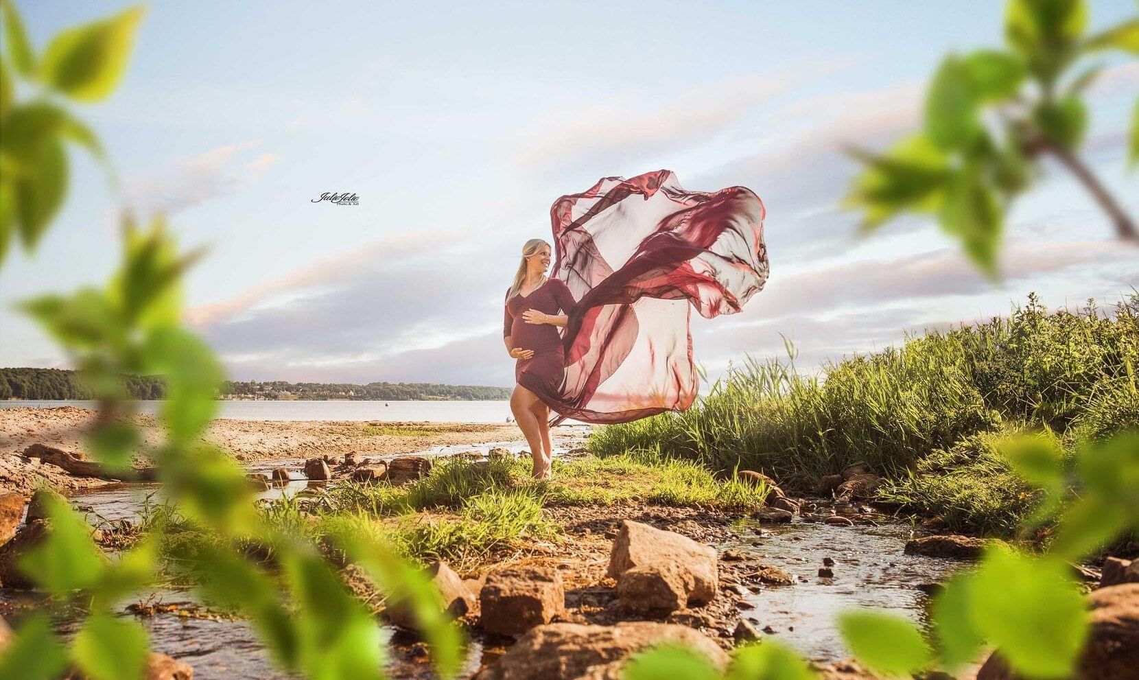 A pregnant woman is standing on a wild beach in a burgundy red dress with a long, fluttering train.