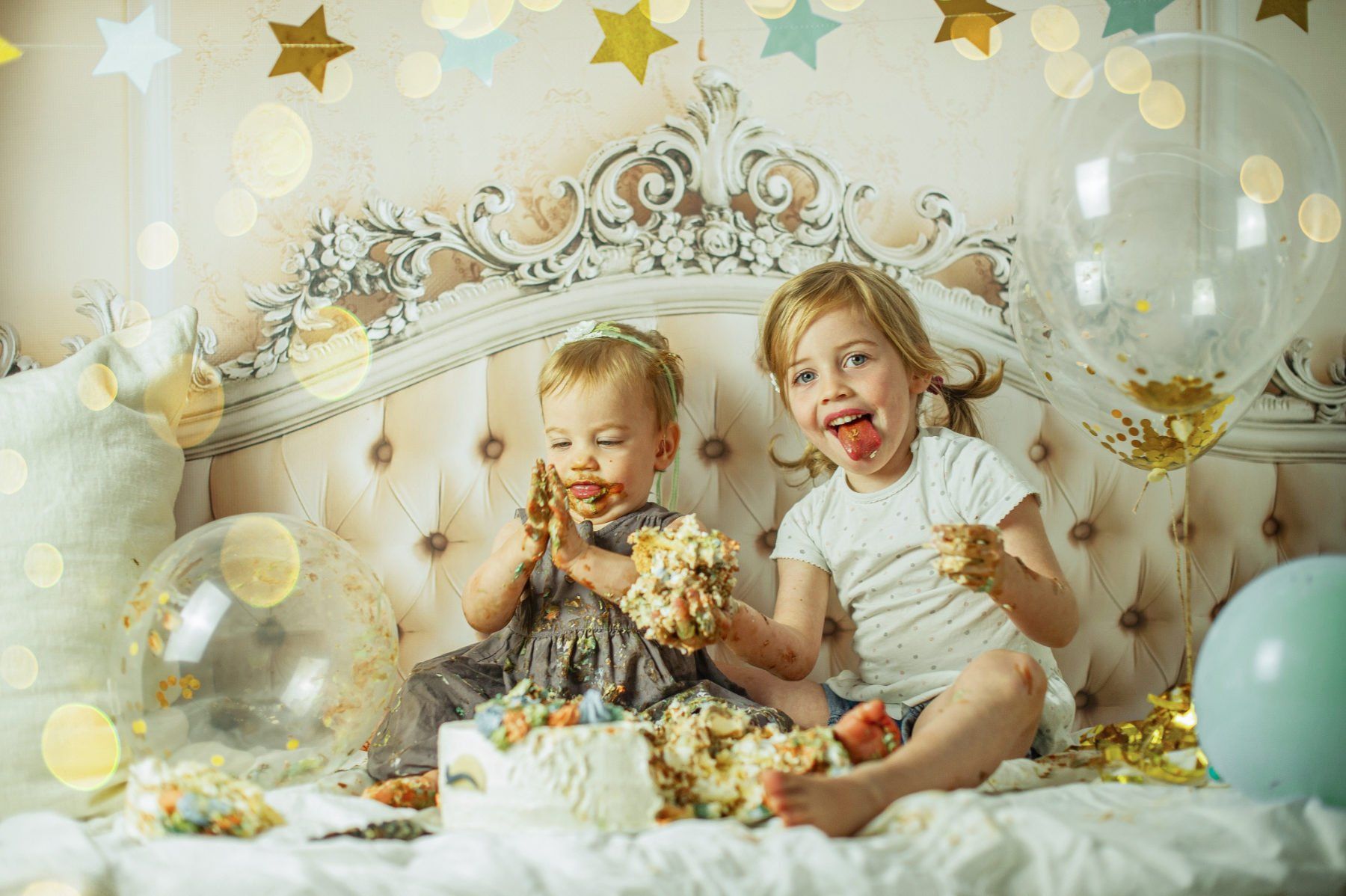 A one year old girl and the older sister are sitting on the bed with the birthday cake in their hands, face and bare feet. Transparent balloons float next to them.