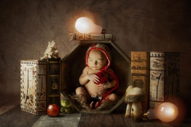 A newborn baby sits in a hexagonal wooden box with a light bulb over it and books for decoration.