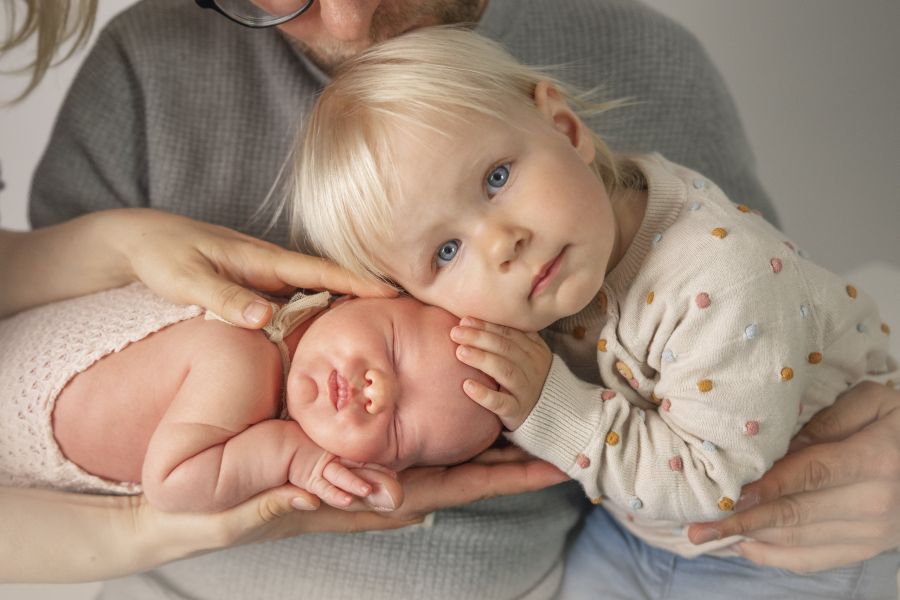 Babyfotografie mit der Familie. Ein Baby liegt schlafend auf dem Unterarm des Vater, ein anderes kleines Kind legt seitlich den Kopf darauf, die Mutter hält ihre Hände unter und über das Baby.