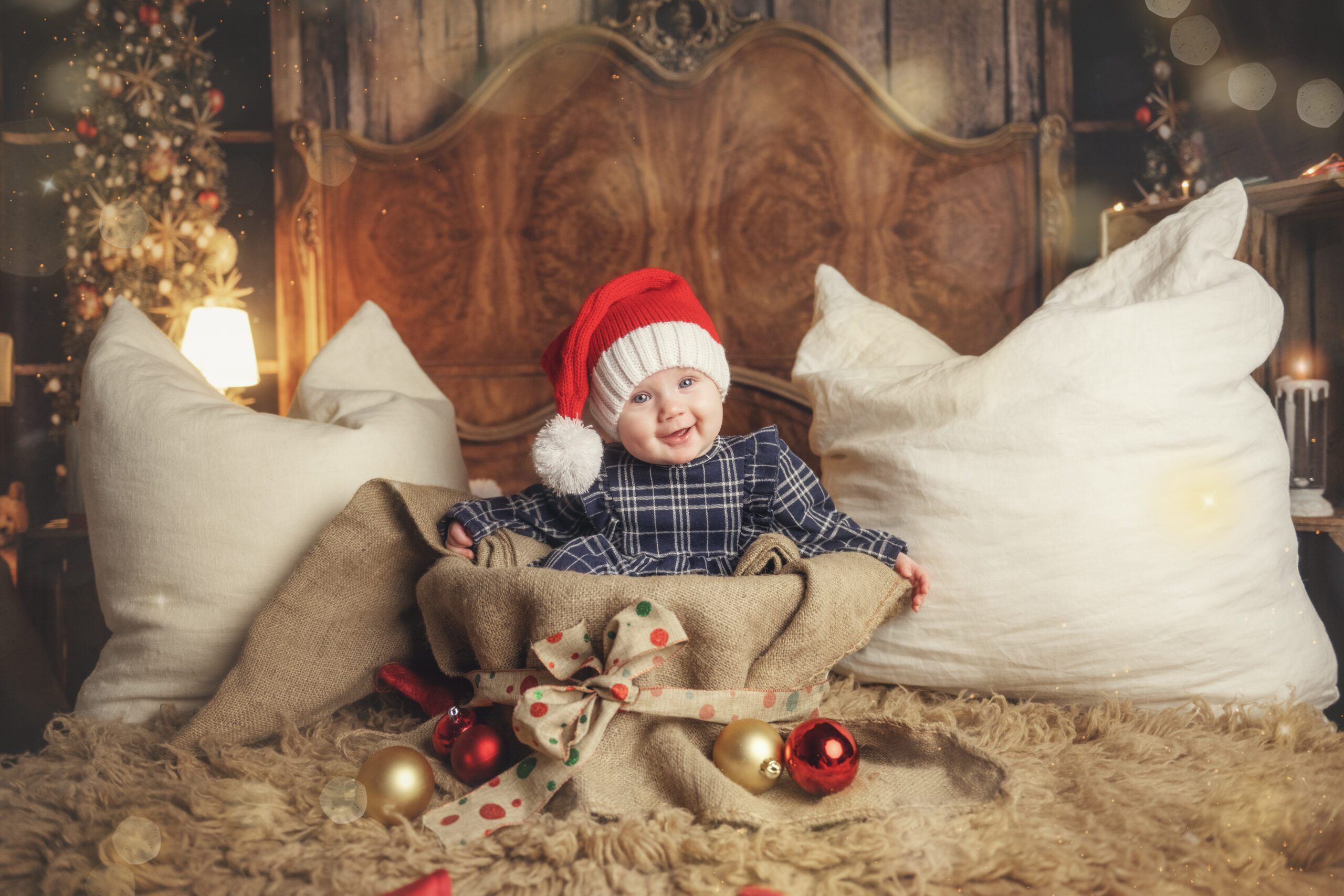 A happy baby wearing a red Christmas hat sits in a sack with a bow on a cozy bed, surrounded by gold and red Christmas baubles.