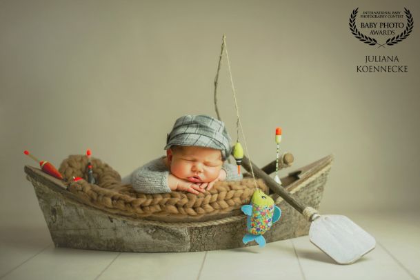 Ein neugeborenes Baby liegt bäuchlings, quer in einem kleinen Ruderboot. Aus dem Boot ragt eine Angel, sowie ein Paddel.