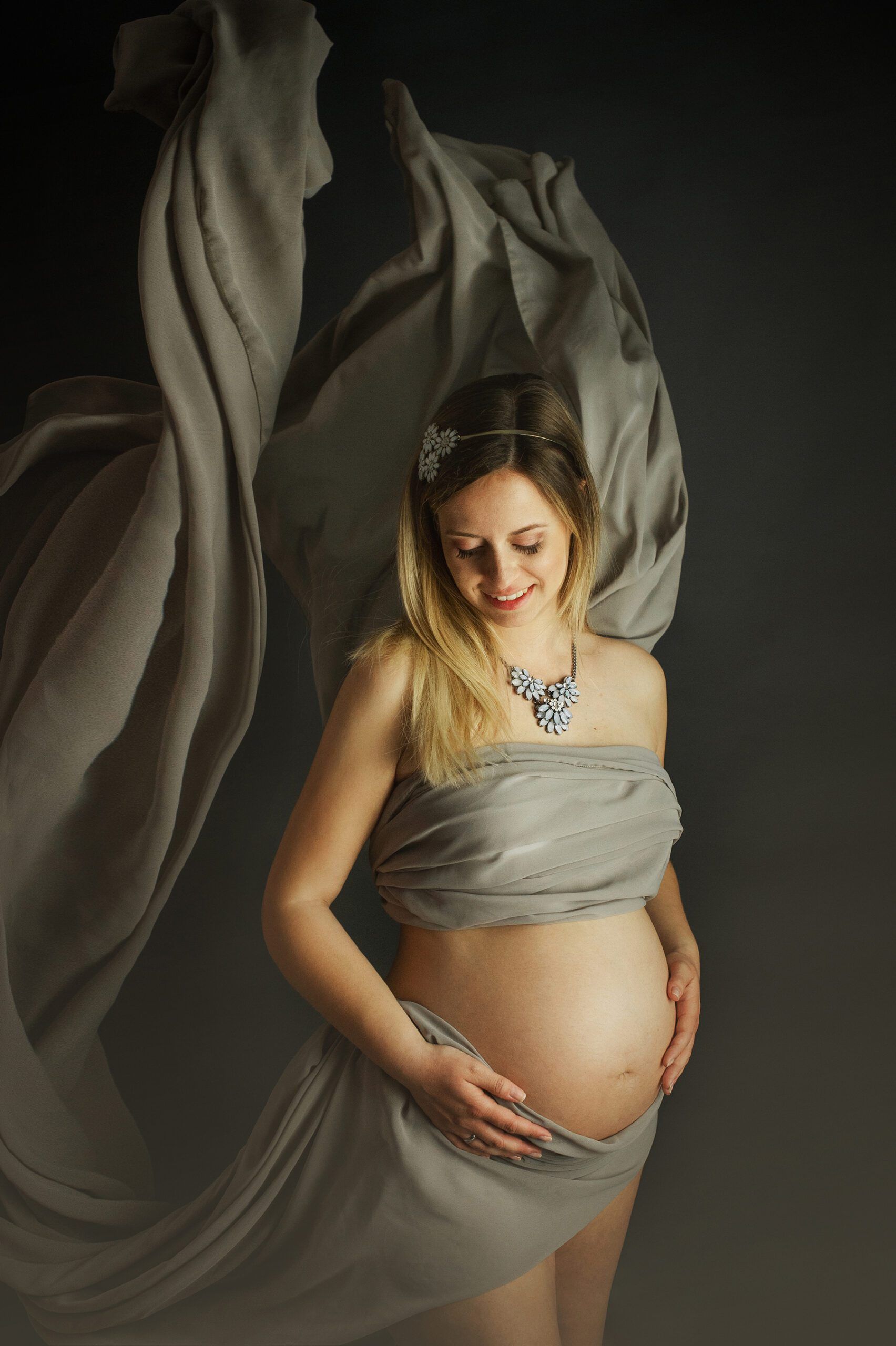 A pregnant woman lightly clothed with a green-gray cloth in front of a dark gray background. The cloths are blowing up in the background. The belly is free and is embraced by it.