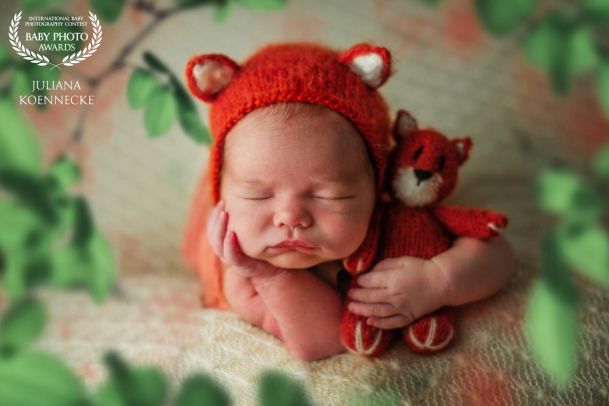 Ein Baby liegt in Bauchlage, den Kopf auf einen Arm gestützt. In der anderen Hand befindet sich ein kleines rotes Kuscheltier passend zur roten Mütze.