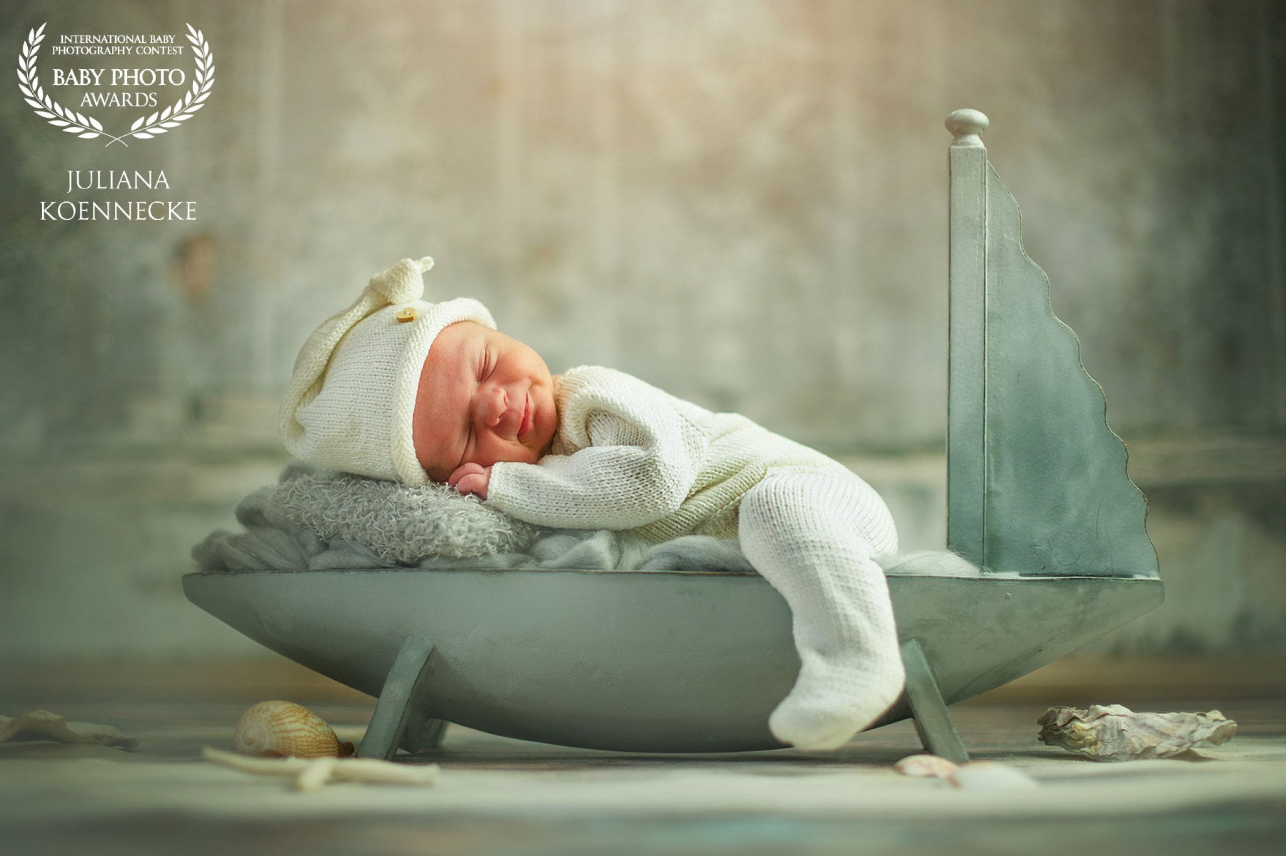 A small decorative sailboat on which a small baby lies on his stomach and sleeps. The baby is smiling and wearing a white knitted romper and matching hat.