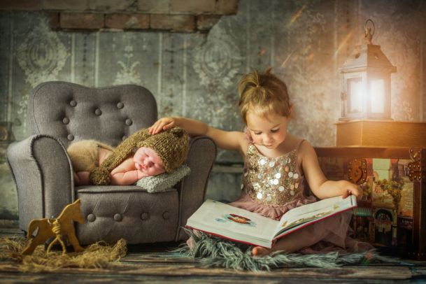 A newborn baby sleeps lying on a small armchair. The sister sits next to it and reads from a large book. A lantern shines in the background.