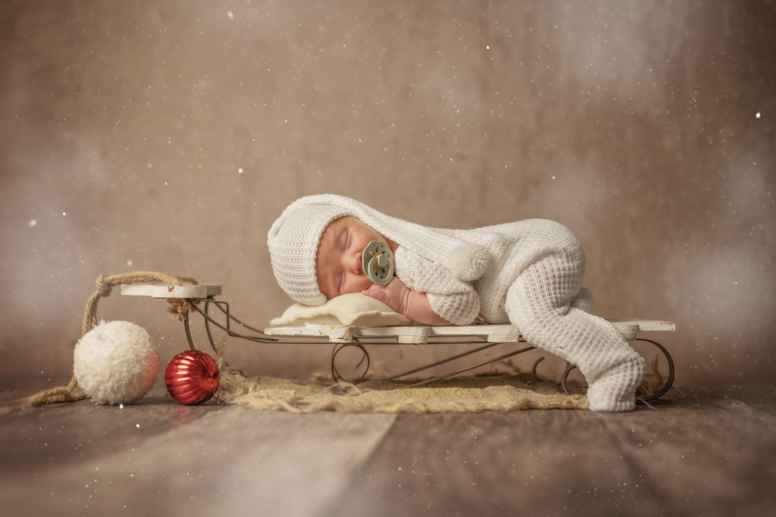 A sleeping newborn in a white knitted jumpsuit with a hood lies on a small wooden sled, surrounded by Christmas decorations and soft light.