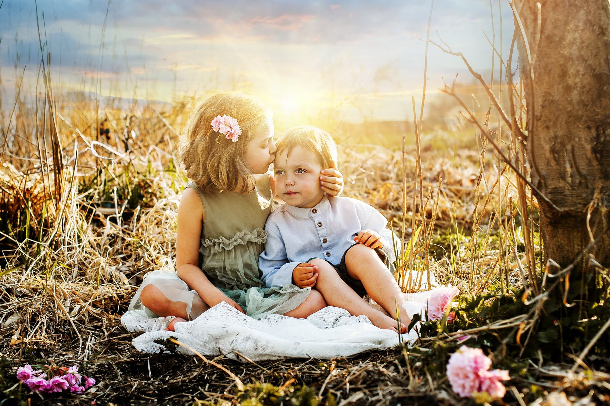 Two children, a little boy and a slightly older sister, are sitting in the dry grass. The girl holds the boy's head and kisses him on the forehead. The sun is setting in the background.