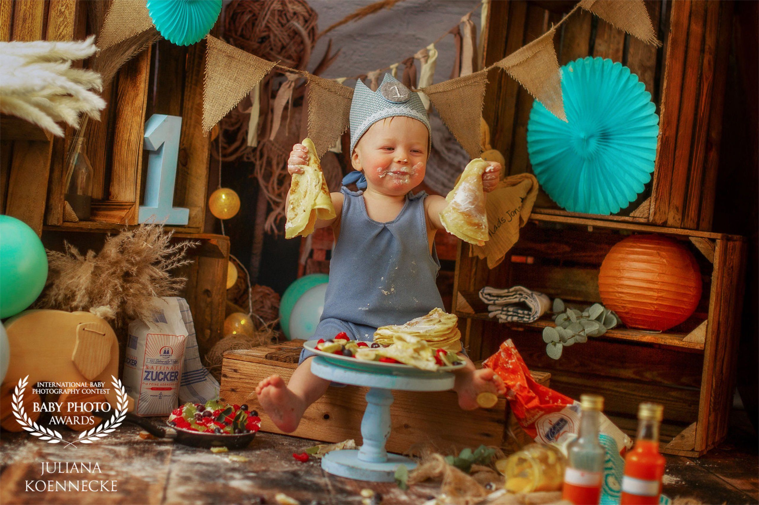 Smash Cake Shooting Hamburg. Ein Baby hält voller Freude mehrere Pfannkuchen in der Hand. Vor ihm ein Teller mit weiteren Pfannkuchen und Früchten. Ein Pancake Shooting als Idee zum Cake Smash.