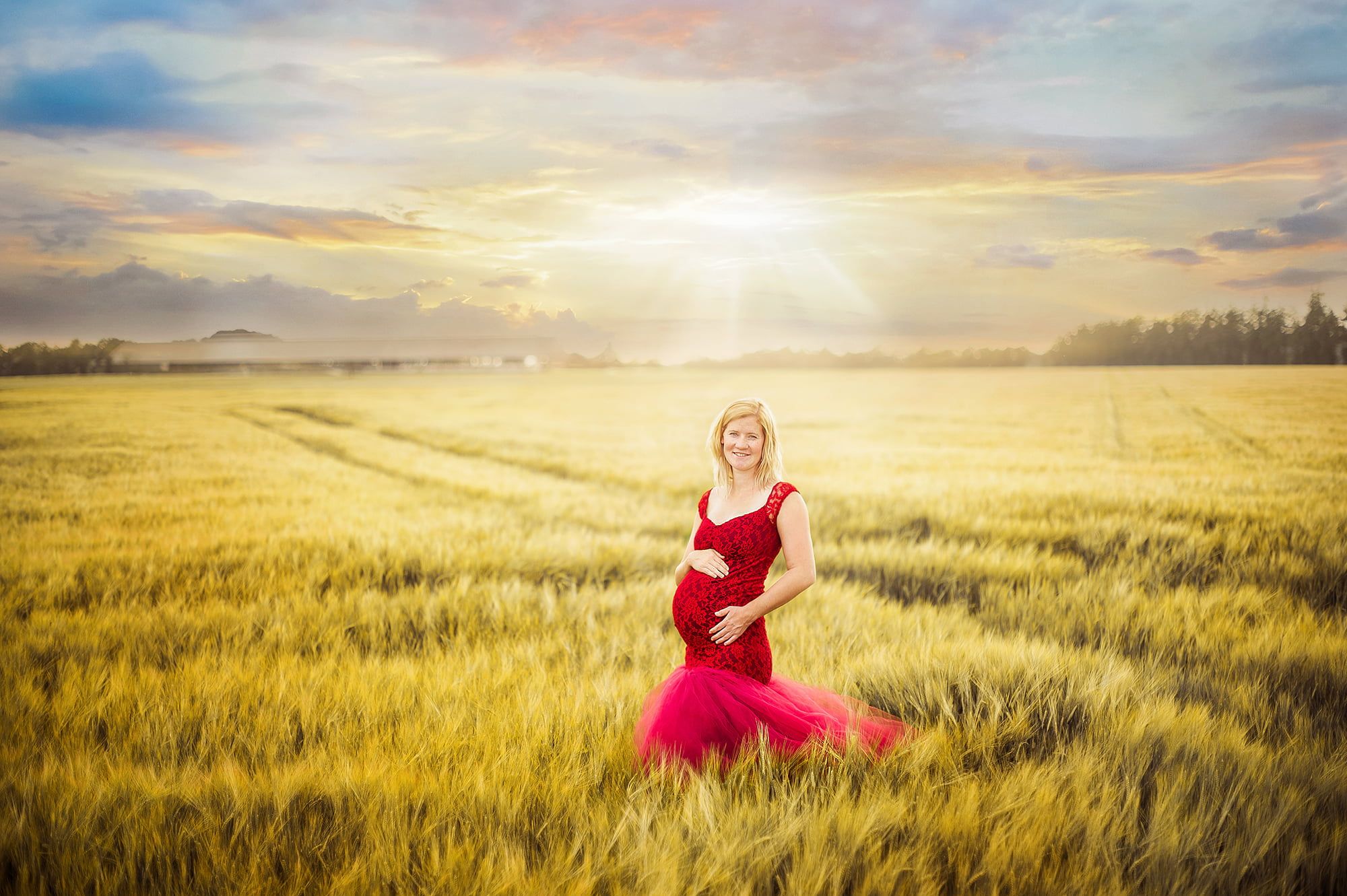 Zu sehen ist ein weites Kornfeld, in dessen Vordergrund eine schwangere Frau mit rotem Trägerkleid steht. Ihre Hände umschließen ihren Bauch, sie lächelt in die Kamera. Im Hintergrund ist der Sonnenaufgang zu sehen.