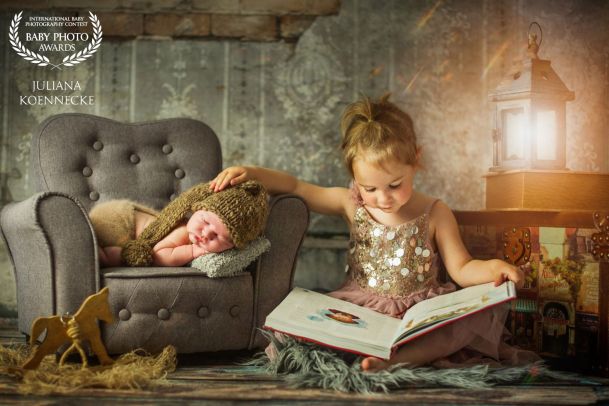 Ein neugeborenes Baby liegt schlafend auf einem Sessel. Daneben sitzt die ältere Schwester und hat ein großes aufgeschlagenes Buch auf Ihren Schultern. Im Hintergrund leuchtet eine Laterne.