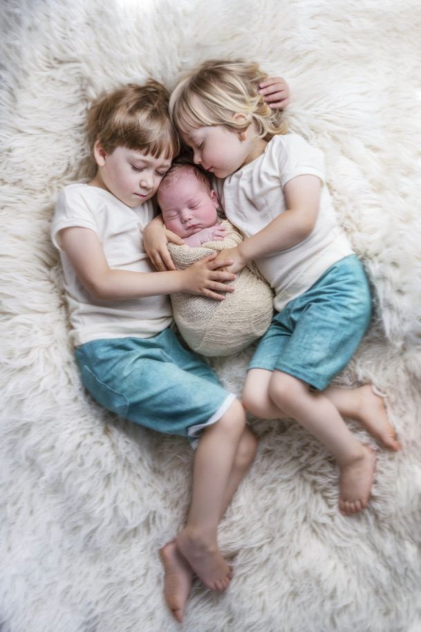 Zwei Kinder liegen eng aneinander gekuschelt, in der Mitte zwischen ihnen liegt, eingewickelt ein Neugeborenes Baby.