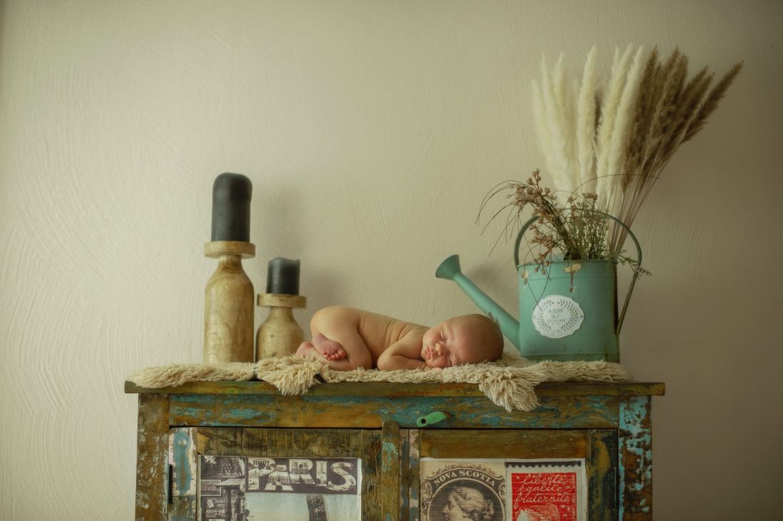 Ein Baby liebt auf einer Kommode auf dem Bauch und schläft. Rechts und links stehe eine grüne Gießkanne und zwei Kerzen.