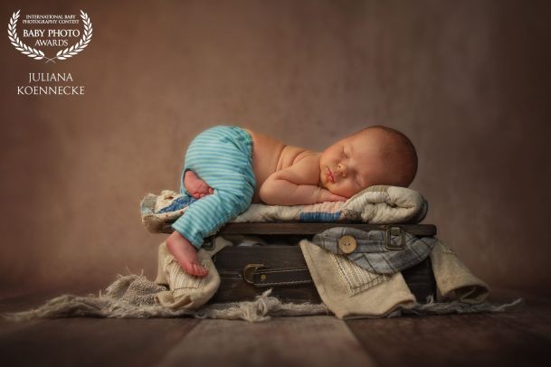 Ein Baby schläft in Bauchlage auf einem kleinen Koffer. Ein Beinchen hängt am Koffer herunter. Verschiedene Kleidungsstücke ragen aus dem Koffer.