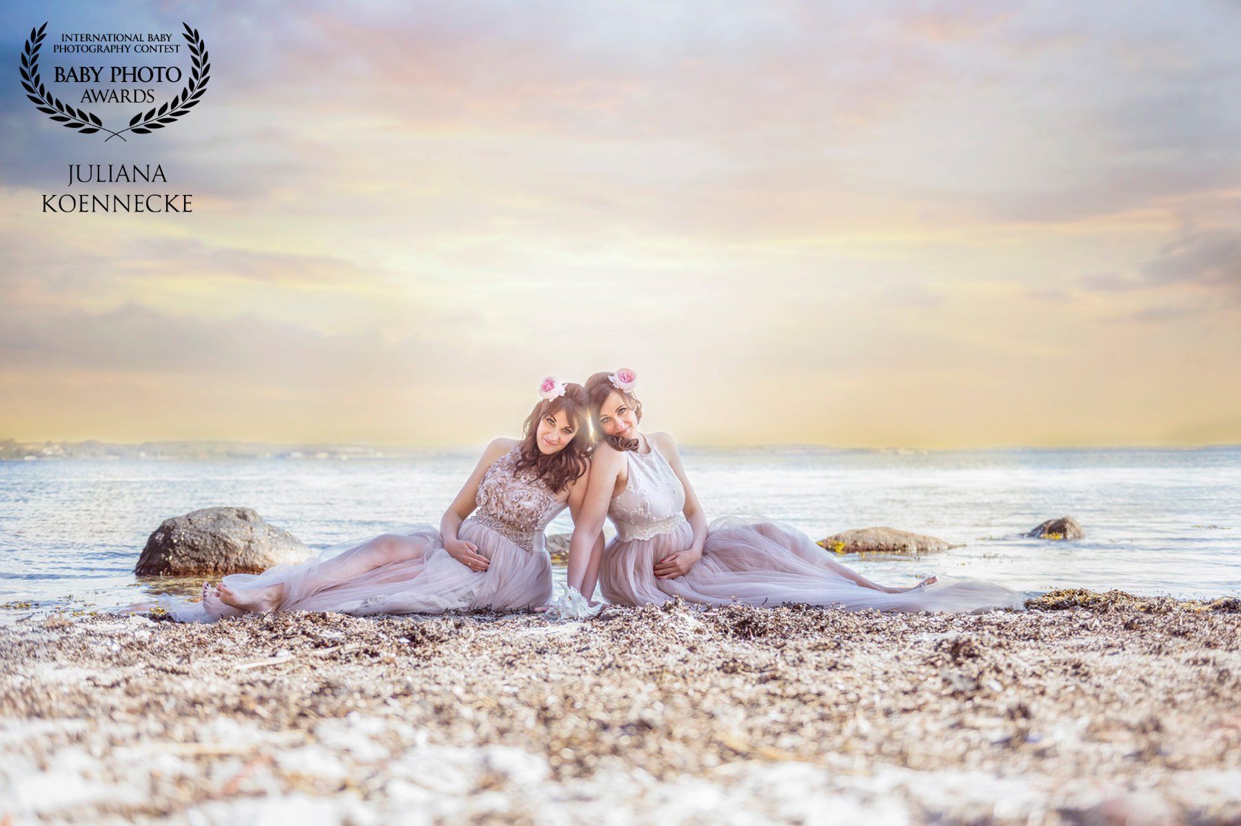 Babybauch Shooting mit schwangeren Zwillingen am Strand. Das Bild zeigt zwei schwangere Frauen, die aneinander gelehnt am Strand liegen. Im Hintergrund das Meer.