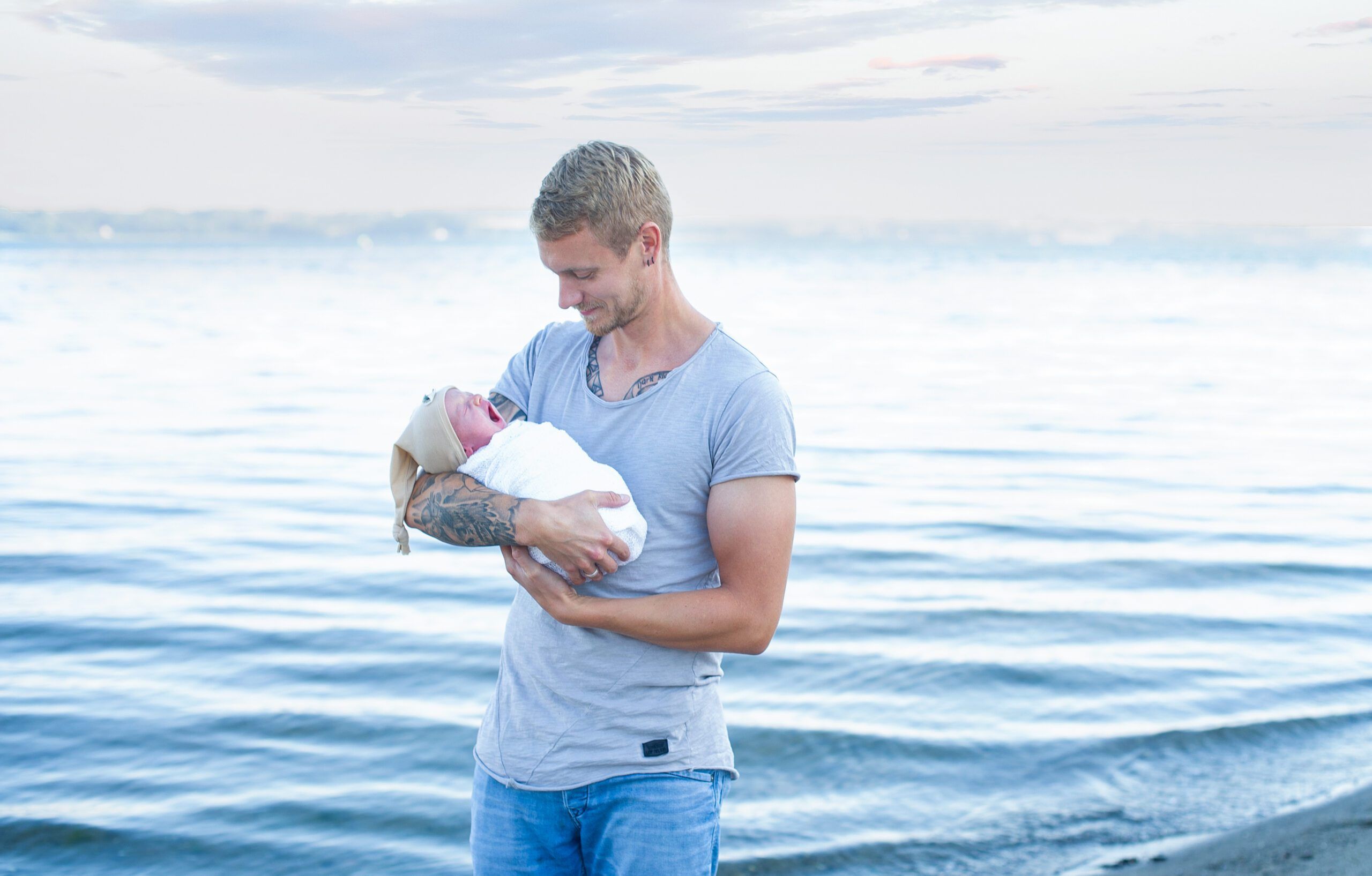 A father is standing on the beach with the newborn baby in his arms. The baby yawns, the father looks at it. The calm sea can be seen in the background.