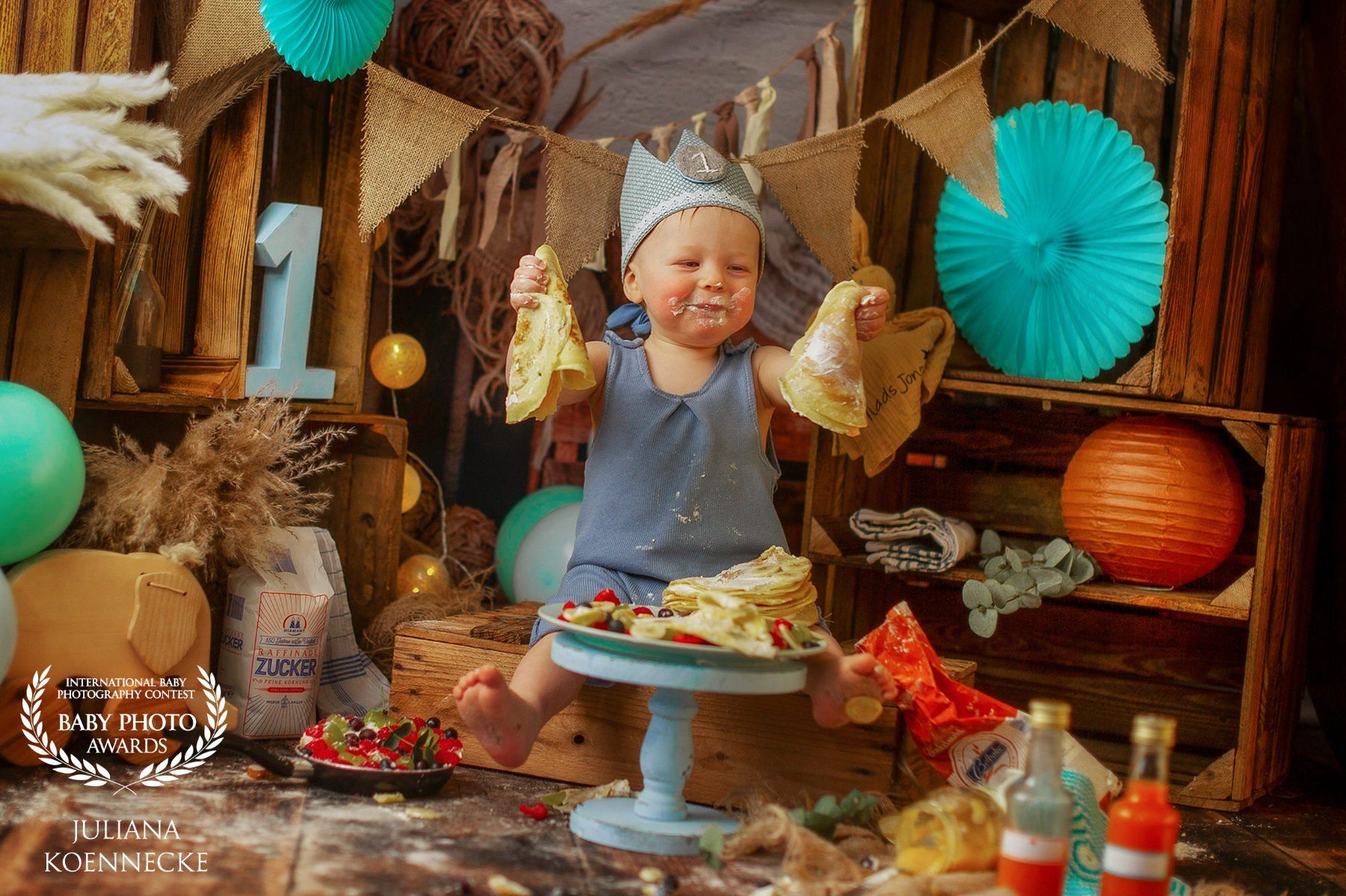 A one year old boy sits beaming in front of a small table covered with fresh pancakes. He holds a pancake dangling in both his right and left hand. The background is decorated with lanterns and natural-colored pennants.