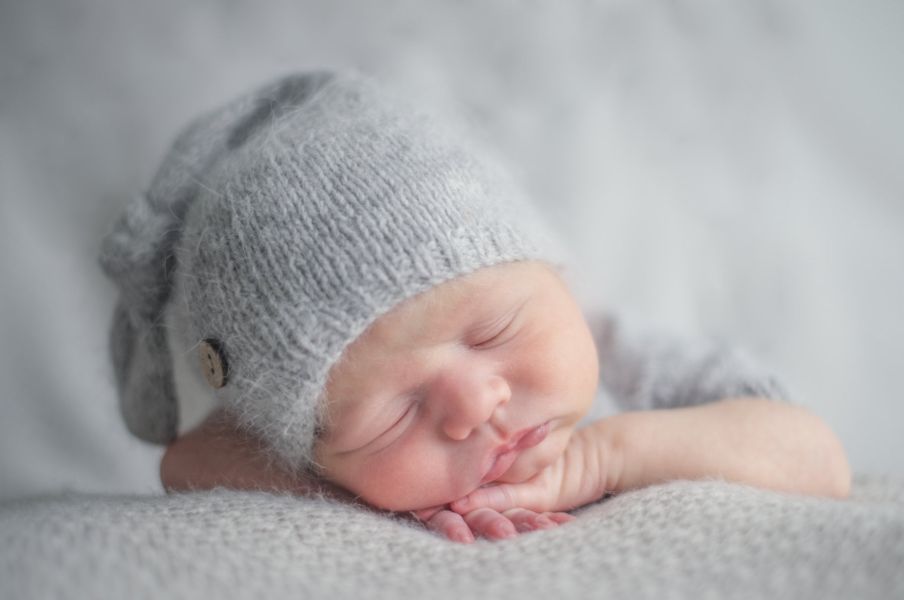 Ein Babyfotoshooting zeigt das Neugeborene auf dem Bauch liegend, mit dem Gesicht zum Betrachter. Es trägt eine kleine Mütze und schläft friedlich auf den Händchen.