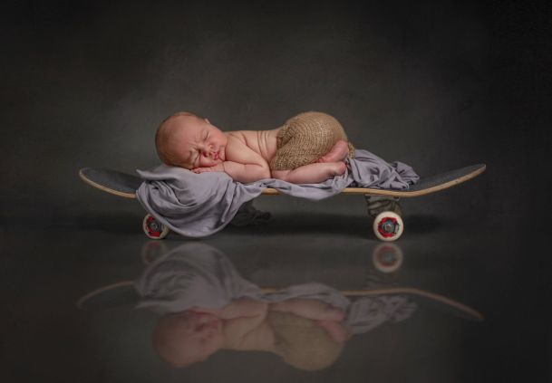 Ein Neugeborenes liegt schlafend auf einem Skateboard. Es wirkt als ob das Skateboard auf einer dünnen Wasserschicht steht.