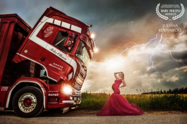 Eine hochschwangere Frau in rotem, langem Kleid steht auf einem Feldweg. Direkt vor ihr steht ein großer, roter LKW mit nach vorne geklapptem Führerhaus. Im Hintergrund ist der Himmel mit Wolken und Blitzen zu sehen.