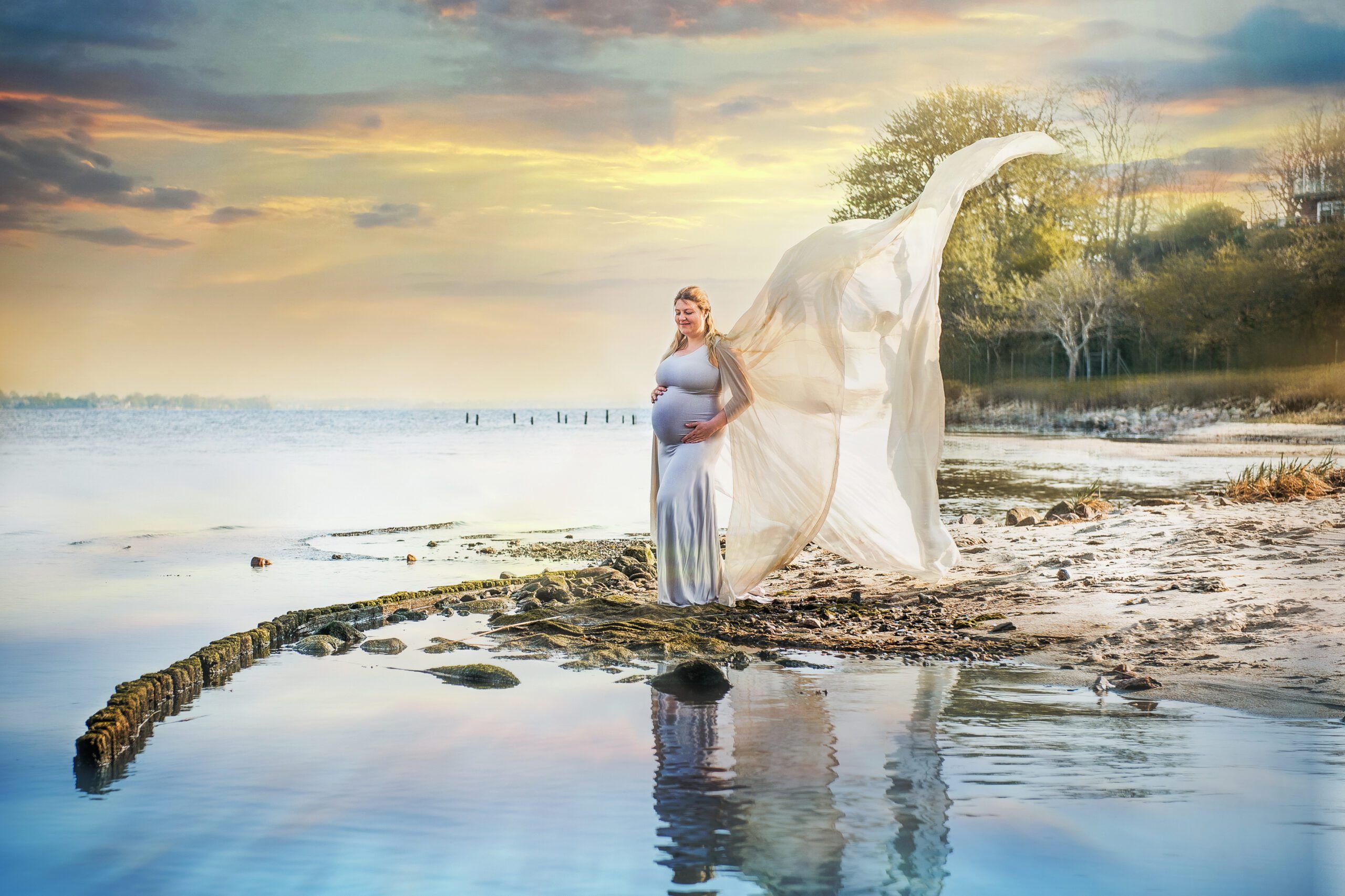 A pregnant woman stands on a rough beach. In front of her and behind her the calm sea. A hill can be seen to the right of her. The train of the dress is blowing in the wind.