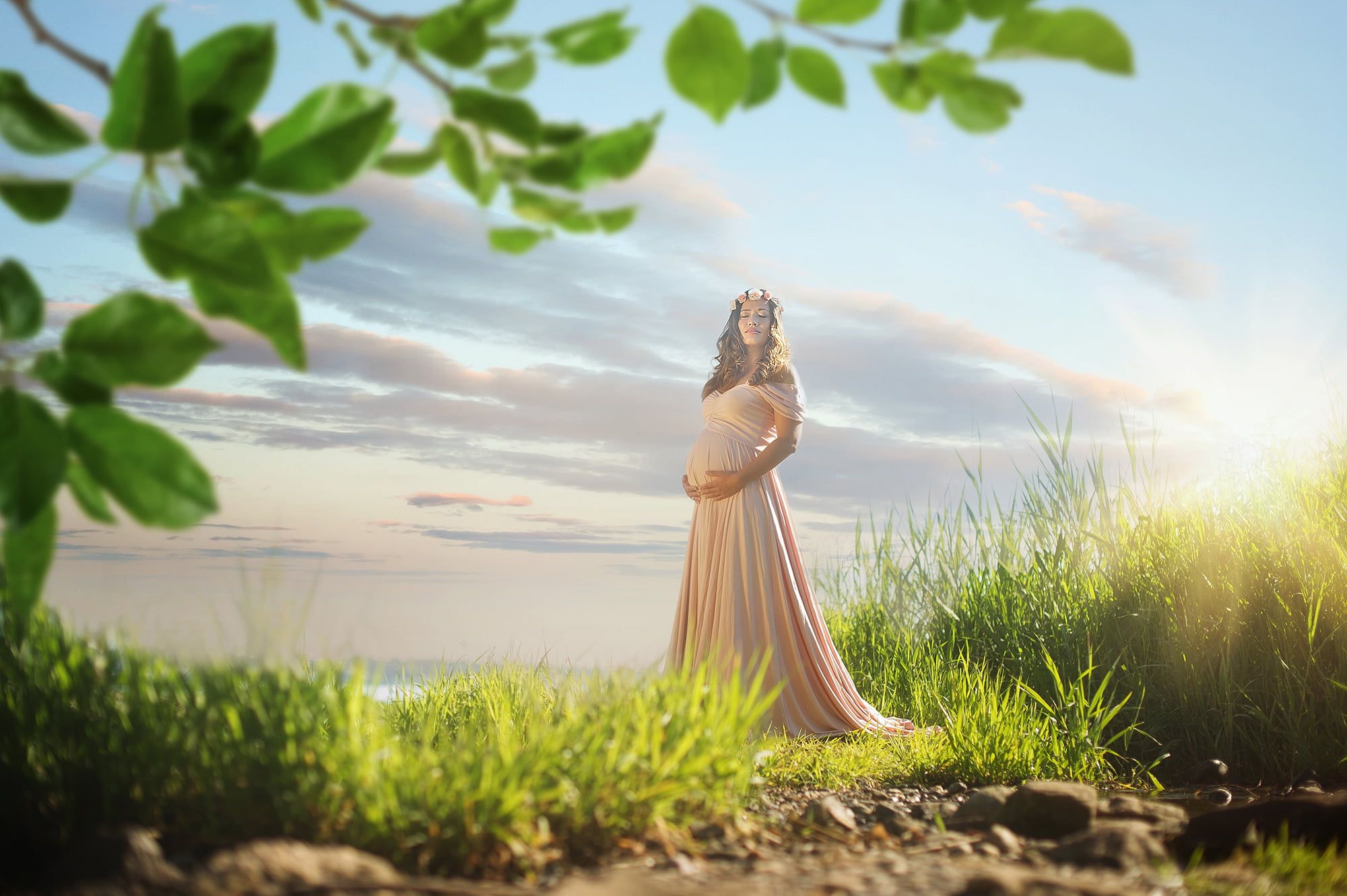 Eine Schwangere junge Frau mit langem pastellfarbenen Kleid steht zwischen Gräsern, im Hintergrund ist das Meer erkennbar. Mein hat den Eindruck als würde sie durch die Büsche im Vordergrund fotografiert werden.