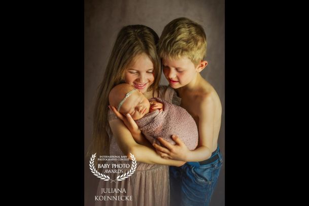 Zwei kleine Geschwister stehen nebeneinander und halten gemeinsam ein Neugeborenes im Arm. Das Baby ist in ein Tuch eingewickelt.